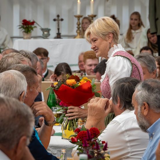 Eine Frau in traditioneller bayerischer Kleidung überreicht freudestrahlend einem Mann, der an einem Tisch sitzt, einen Blumenstrauß. Die Szene spielt in einer überfüllten, festlichen Umgebung mit einem Tisch im Hintergrund, der mit Blumen und Kerzen geschmückt ist.