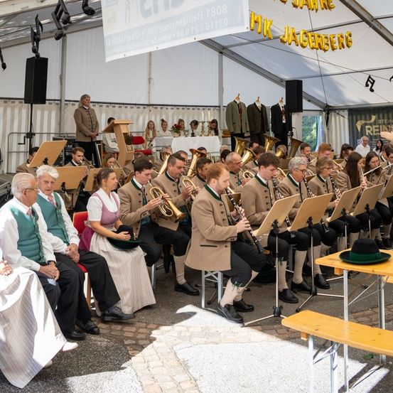 Ein Outdoor-Musikauftritt, eine Band spielt Instrumente wie Trompeten und Klarinetten, mit Zuschauern vor Ort. Ein Banner mit Text hängt von der Zeltdecke ab.