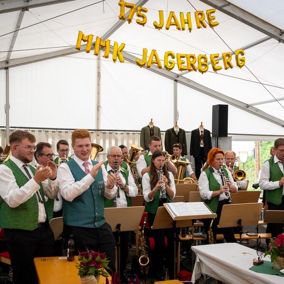 Ein Blasorchester tritt in einem Zelt auf und feiert 175 Jahre MMK Jaegerberg. Sie spielen Instrumente, mit einem Banner im Hintergrund.
