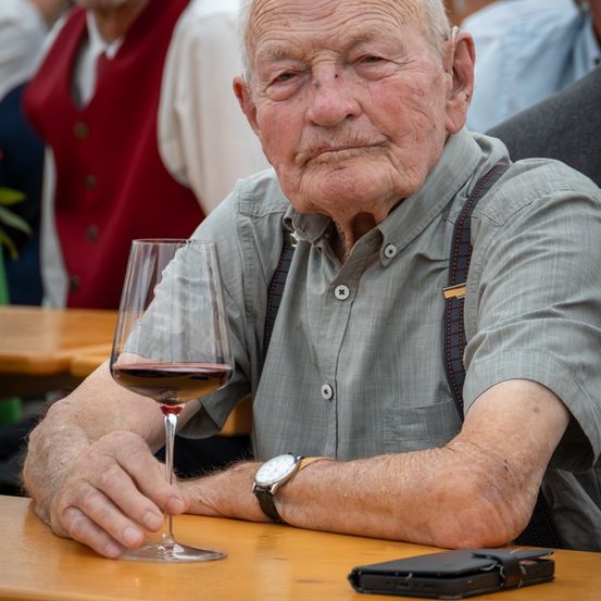 Ein älterer Mann hält ein Glas Rotwein, sitzt an einem Tisch mit einem Handy in der Nähe, trägt eine Hosenträger und eine Uhr. Im Hintergrund sind Menschen um Tische versammelt.