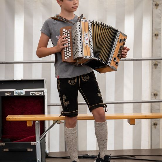 Ein junger Junge steht auf der Bühne und hält ein braunes Akkordeon in der Hand. Er trägt ein graues T-Shirt, schwarze Shorts und graue Socken. Hinter ihm sind eine Bank und ein schwarzer Koffer zu sehen.