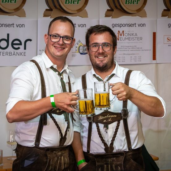 Zwei Männer in traditioneller deutscher Kleidung, mit Trägerhosen und Krawatten, lächeln und halten Bierkrügen hoch. Sie stehen vor einer Wand mit Schildern und Bannern.