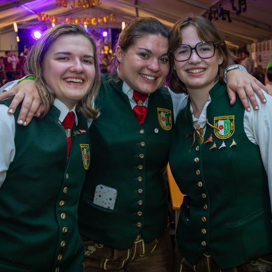 Drei Frauen in grünen Westen und roten Krawatten lächeln in die Kamera, wahrscheinlich bei einem Festival. Sie haben Anstecknadeln und Abzeichen auf ihren Westen. Hinter ihnen sind verschwommene Figuren zu sehen, möglicherweise bei einem Konzert.