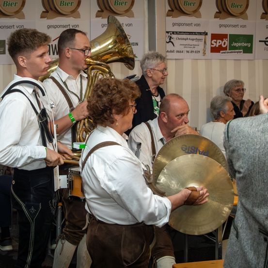 Eine Gruppe von Musikern tritt bei einer Veranstaltung im Freien auf. Dazu gehören ein Tubaspieler, ein Beckenspieler und andere. Hinter ihnen beobachten einige Menschen. Im Hintergrund befinden sich Bierfässer und Banner.