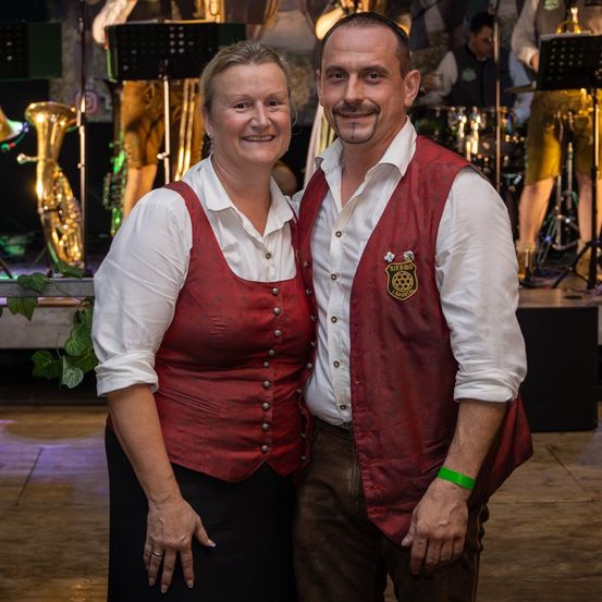 Ein lächelndes Paar in traditioneller Kleidung steht auf einer Bühne, mit einer Musikband im Hintergrund.