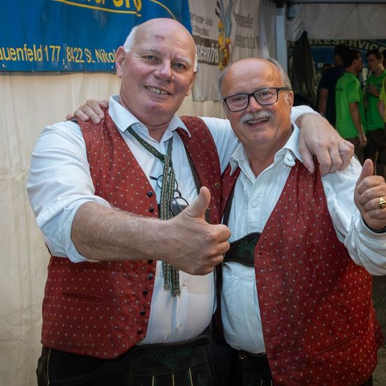 Zwei Männer in traditioneller deutscher Kleidung mit roten Westen und Krawatten geben ein Daumen hoch, lächeln und stehen bei einem Outdoor-Event zusammen.