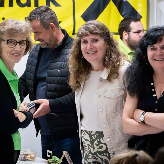 Vier Erwachsene stehen in einer Gruppe. Die Frau auf der linken Seite hält eine Brieftasche und lächelt. Die mittlere Frau trägt ein weißes Oberteil und eine gemusterte Hose. Der Mann hinter ihr trägt eine schwarze Jacke.