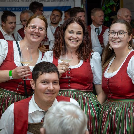 Vier Personen in traditioneller deutscher Kleidung lächeln und halten Weingläser. Hinter ihnen stehen mehrere Personen, einige in ähnlicher Kleidung, und unterhalten sich.