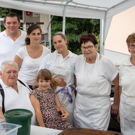 Eine Gruppe von Menschen, einschließlich Erwachsener und Kindern, steht unter einem Zelt. Sie tragen weiße Schürzen. Ein Mädchen in einem Leoparden-Kleid sitzt vor einem Holztisch.