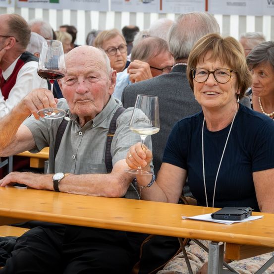 Ein älteres Paar sitzt an einem Tisch und hält Weingläser mit rotem und weißem Wein. Sie sind von anderen Menschen bei einer Veranstaltung im Freien umgeben.