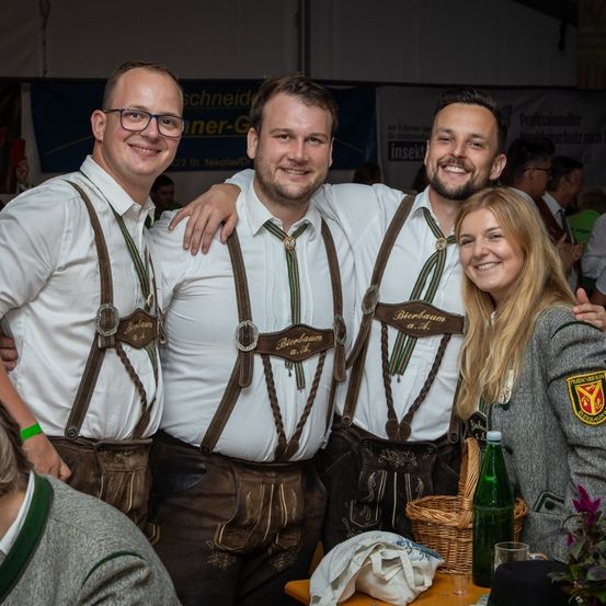 Vier Personen stehen für ein Foto bei einem Festival zusammen, drei Männer in traditioneller bayerischer Kleidung und eine Frau in einer grauen Jacke mit einem Aufnäher. Sie alle lächeln. Dahinter steht ein Tisch mit einem Korb, einer Flasche und einem Glas.