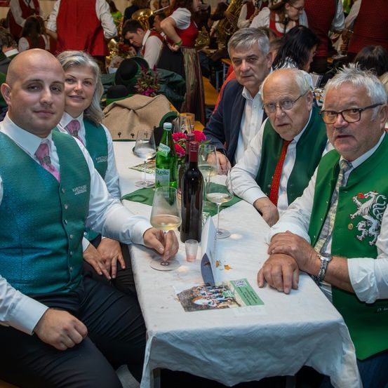 Eine Gruppe von Erwachsenen in grünen Westen und Krawatten sitzt an einem Tisch mit Weingläsern, Flaschen und einer Menükarte. Sie nehmen möglicherweise an einer Party teil.