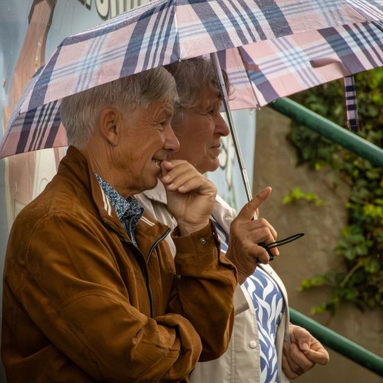 Zwei ältere Menschen stehen unter einem karierten Regenschirm, wahrscheinlich im Regen. Der Mann lächelt und hält sein Kinn, während die Frau etwas ansieht.