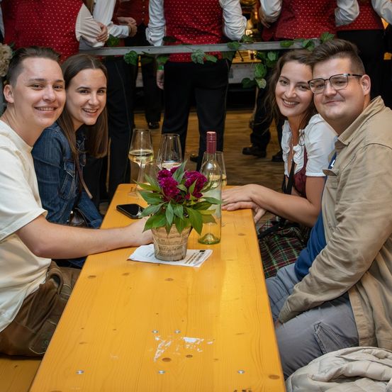 Vier Personen sitzen zusammen an einem Tisch und lächeln für ein Foto. Sie sind umgeben von Weingläsern und einer Topfpflanze. Im Hintergrund stehen andere in traditioneller Kleidung hinter einem Geländer.