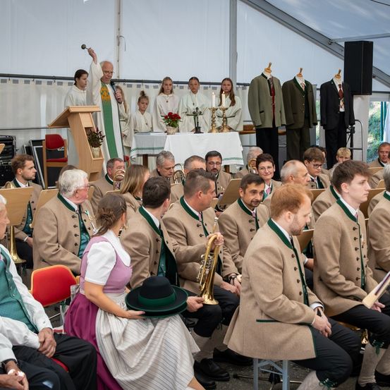 Eine religiöse Zeremonie findet in einem Zelt statt. Ein Priester spricht und hält ein Mikrofon. Chor und Orchester sitzen vor ihm. Das Publikum sitzt auf Stühlen, einige stehen. Der Bereich ist mit traditioneller Kleidung und Kerzen geschmückt.