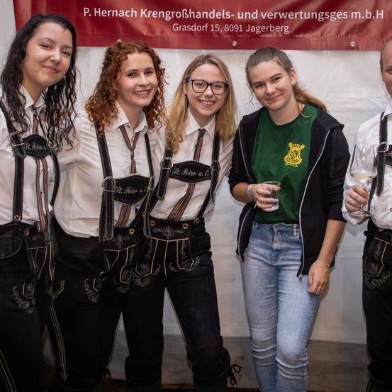 Fünf Frauen posieren für ein Foto vor einem Banner. Vier tragen traditionelle deutsche Kleidung mit Lederhosen und eine trägt ein grünes Shirt und Jeans. Sie lächeln und halten Gläser mit Wein in der Hand.