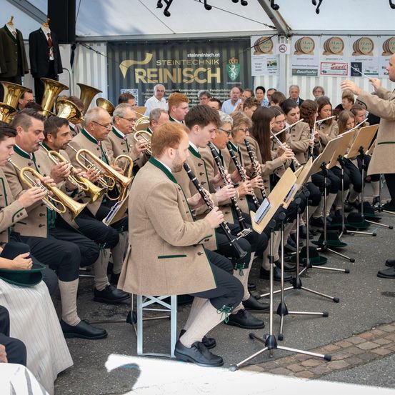 Ein Blasorchester mit Trompeten und Klarinetten spielt im Freien, mit einem Dirigenten an der Spitze und Zuschauern im Hintergrund.