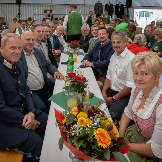 Eine Gruppe von Menschen sitzt an einem langen Tisch mit Blumen und lächelt für ein Foto. Die Umgebung scheint ein Bankettsaal zu sein, mit Musikern im Hintergrund.