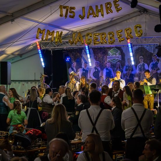 Eine Gruppe Musiker tritt auf einer Bühne unter einem Zelt auf, das mit 175 Jahre MK Jagerberg gekennzeichnet ist. Sie spielen verschiedene Instrumente, während Menschen davor zusehen.