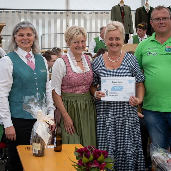Vier Erwachsene stehen lächelnd vor einem Tisch mit Blumen und Geschenken. Eine Frau in einem blauen Kleid hält ein Zertifikat für 300 Euro. Sie befinden sich in einer festlichen, möglicherweise im Freien stattfindenden Veranstaltung.