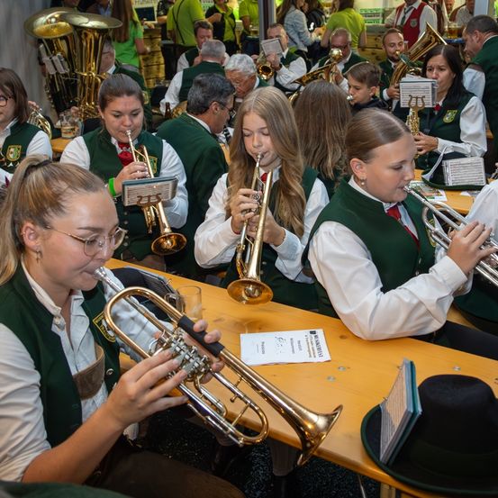 Eine Gruppe von Musikern, darunter junge Frauen, spielen Blechblasinstrumente, wahrscheinlich in einem Musiksaal, mit Musikblättern und Hüten in der Nähe.