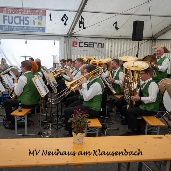 Eine Blechbläsergruppe spielt in einem Zelt. Die Musiker tragen grüne Westen. Ein Banner mit dem Wort 'FUCHS' ist an der Wand angebracht. Vor ihnen steht ein hölzerner Tisch mit einer Pflanze.