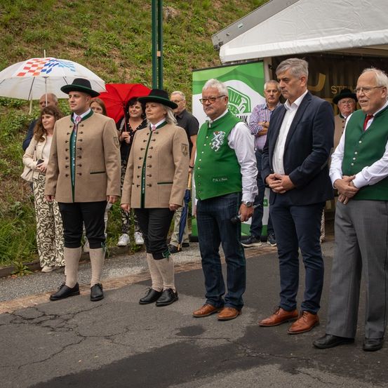 Eine Gruppe von Menschen in traditioneller bayerischer Kleidung steht auf einer Straße, ein Mann hält ein Mikrofon. Hinter ihnen versammeln sich andere, einige halten Regenschirme. Im Hintergrund steht ein Zelt mit einem grünen Banner.
