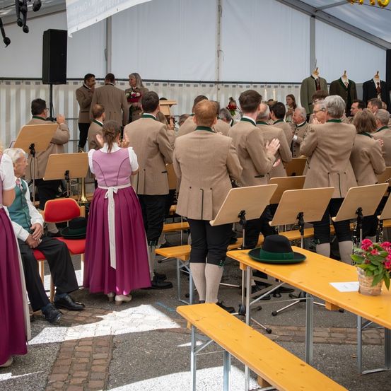In einem Zelt singt ein Chor in traditioneller Kleidung, während Leute auf Stühlen sitzen und ein Tisch mit Hut und Blumen zu sehen ist.