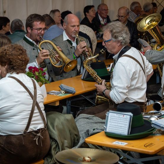 In einem Raum spielen mehrere Musiker Instrumente, darunter Trompete, Saxophon und Tuba. Einige Leute beobachten sie, und auf den Tischen liegen Notenblätter und Blumen.