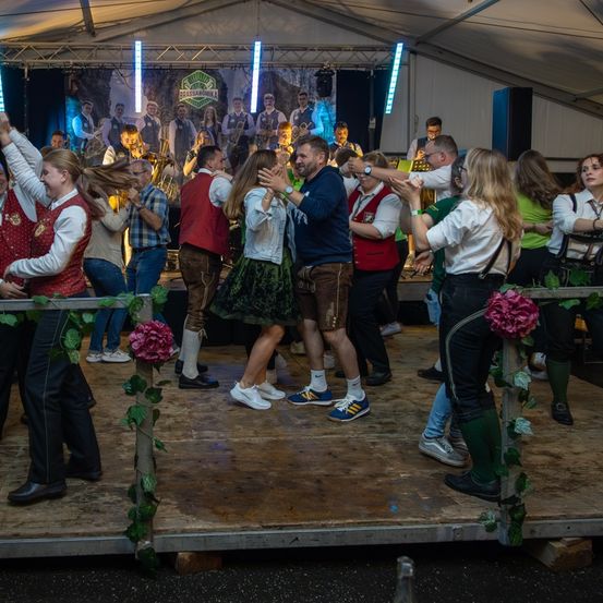 Menschen tanzen auf der Bühne in traditioneller Kleidung unter einem Zelt, während eine Band im Hintergrund spielt. Die Bühne ist mit Blumen geschmückt und es gibt Lichter an der Decke.