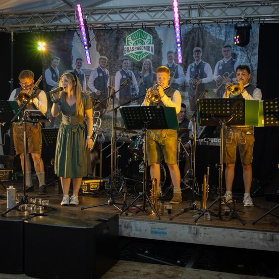 Eine Gruppe von Musikern auf der Bühne spielt mit Trompeten und Posaunen. Ein großer Hintergrund zeigt eine Gruppe von Menschen in traditioneller Kleidung.