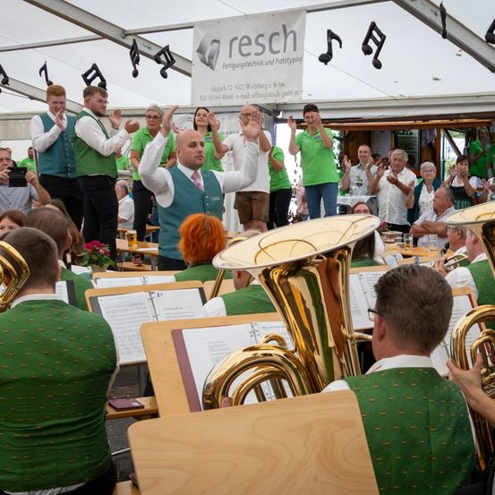 Eine Band spielt unter einem Zelt mit einem Banner. Die Mitglieder in grünen Westen spielen Instrumente und eine Menge schaut zu. Musiknoten hängen über ihnen.