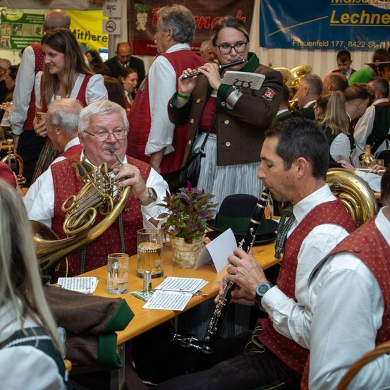 Eine Gruppe von Menschen spielt Musikinstrumente in einem überfüllten Raum. Ein Mann spielt Trompete, ein anderer Mann spielt Klarinette und eine Frau spielt Flöte. Hinter ihnen sitzen und stehen Menschen. Auf dem Tisch steht eine Pflanze in einem Topf.