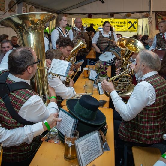 Eine Gruppe von Musikern in traditioneller Kleidung spielt Blechblasinstrumente bei einer Veranstaltung im Freien. Die Szene zeigt sitzende und stehende Personen, mit sichtbaren Musikinstrumenten und Notenblättern.