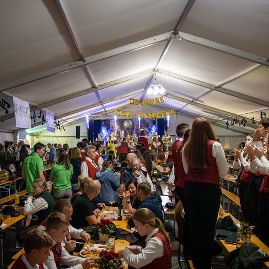 Eine Gruppe von Menschen versammelt sich in einem Zelt. Einige sitzen an Tischen, andere stehen und klatschen. Das Zelt ist mit Bannern geschmückt und im Hintergrund ist eine Bühne mit einer Band zu sehen.