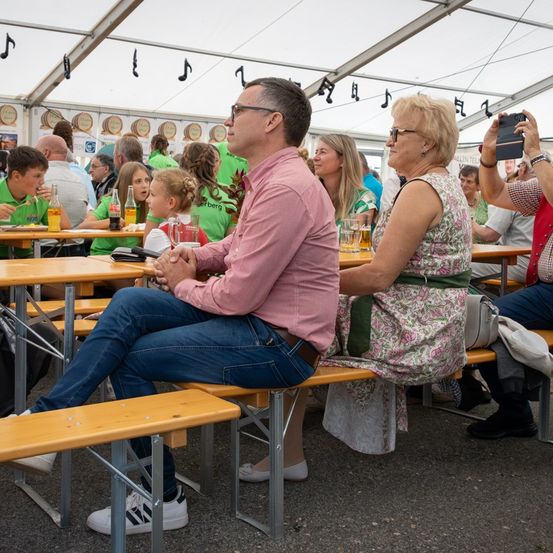 Menschen sitzen auf Bänken in einem Zelt. Ein Mann und eine Frau sitzen nebeneinander. Hinter ihnen führen andere Gespräche. Die Szene umfasst Tische mit Getränken und Essen.