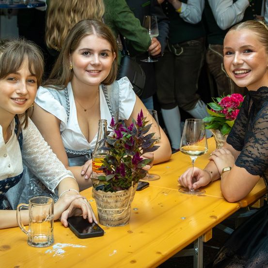Drei Frauen sitzen an einem Tisch, lächeln und posieren für ein Foto. Sie haben Weingläser, einen Topf mit Pflanzen und ein Handy auf dem Tisch. Hinter ihnen stehen Menschen und tragen traditionelle Kleidung.