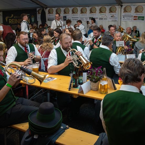 Eine Gruppe von Menschen in grünen Westen und Hüten spielt in einem Zelt. Sie spielen Musikinstrumente wie Trompeten, Posaunen und Klarinetten. Auf dem Tisch vor ihnen stehen mehrere Flaschen und Gläser.