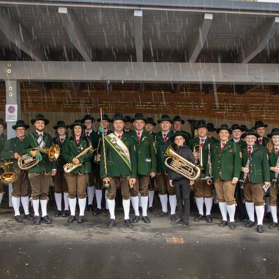 Eine Blaskapelle in grünen Uniformen posiert für ein Foto im Regen. Jedes Mitglied hält ein Blechblasinstrument.