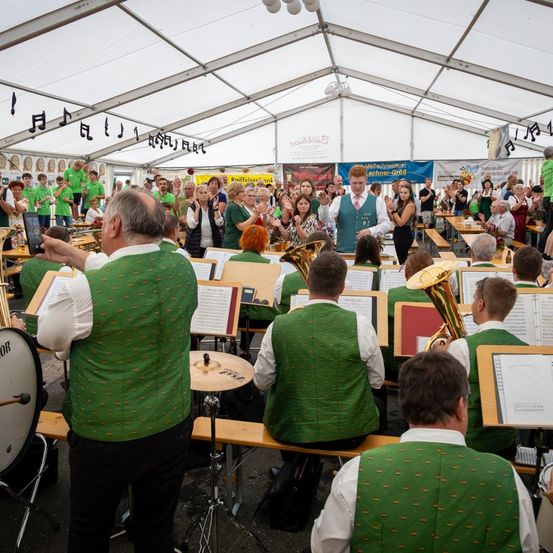 Eine Band spielt Musik unter einem Zelt, mit Musikern in grünen Westen und einem Publikum, das klatscht. Musiknoten sind an der Zeltdecke zu sehen, und die Szene deutet auf ein festliches oder kulturelles Ereignis hin.