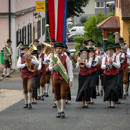 Eine Blaskapelle mit Blech- und Holzblasinstrumenten zieht in traditioneller Kleidung die Straße entlang. Gebäude und Bäume säumen die Straße, im Hintergrund befindet sich ein Banner.