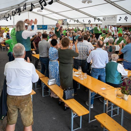 Eine Gruppe von Menschen in Grün und Weiß klatscht in einem Zelt. Musiknoten und ein Banner mit dem Wort reschi hängen von der Decke. Sie sitzen an Tischen mit Weingläsern und einer Handtasche.