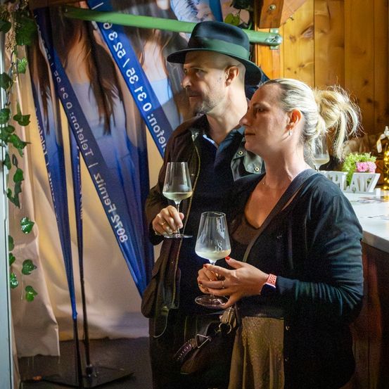 Ein Mann und eine Frau in einer Bar, der Mann trägt einen Hut, beide halten Weingläser. Ein blauer Banner mit Text dahinter.