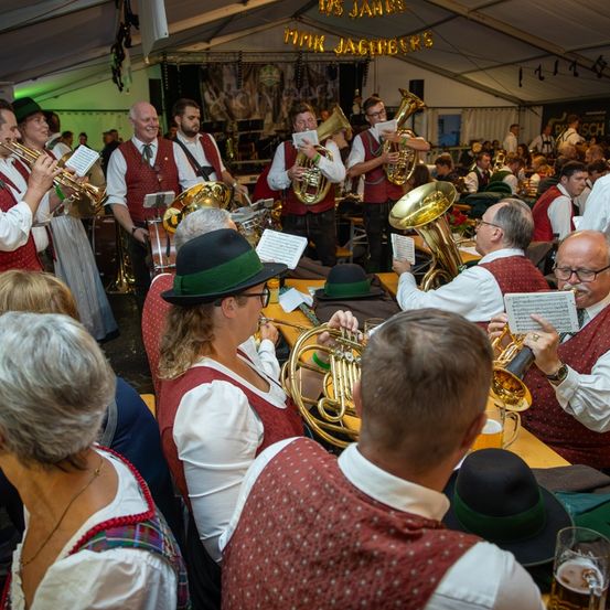 Eine Gruppe von Musikern, einige spielen Blechblasinstrumente, tritt in einem Zelt mit festlicher Atmosphäre auf. Einige Mitglieder tragen traditionelle Kostüme.