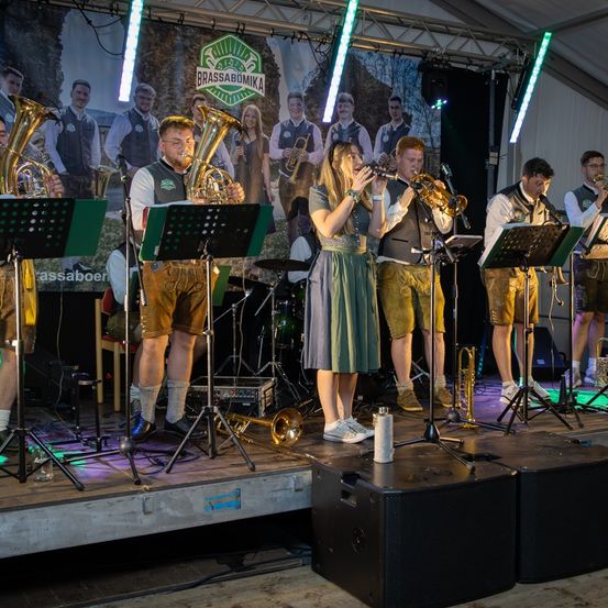 Eine Brassband spielt auf der Bühne mit verschiedenen Instrumenten wie Tubas, Trompeten und Posaunen. Ein großes Banner ist im Hintergrund zu sehen.