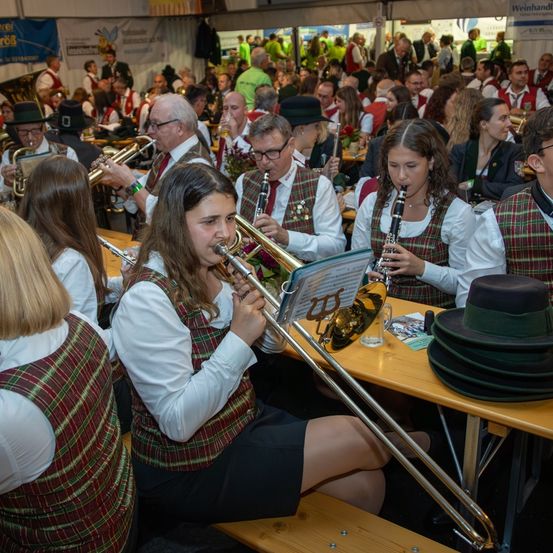 Eine Gruppe von Musikern, hauptsächlich junge Frauen, spielen verschiedene Instrumente bei einer festlichen Veranstaltung. Die Instrumente umfassen Posaunen, Klarinetten und Trompeten. Das Publikum sitzt und beobachtet die Aufführung.