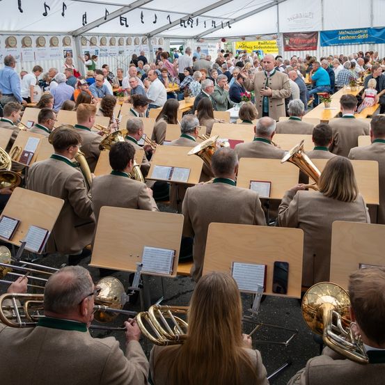 Ein großes Blasorchester spielt unter einem Zelt, während das Publikum auf Bänken und Tischen sitzt. Die Musiker spielen verschiedene Instrumente, darunter Tubas und Posaunen. Das Publikum scheint die Aufführung zu genießen.