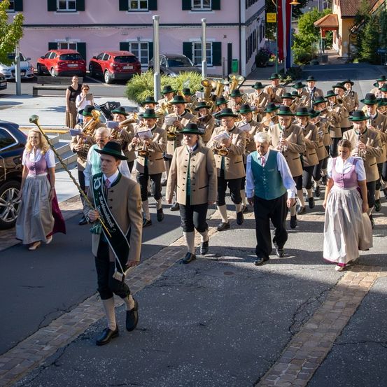 Eine Blaskapelle mit Blech- und Holzblasinstrumenten geht auf einer Straße. Sie tragen traditionelle Kleidung. Autos sind am Straßenrand geparkt.