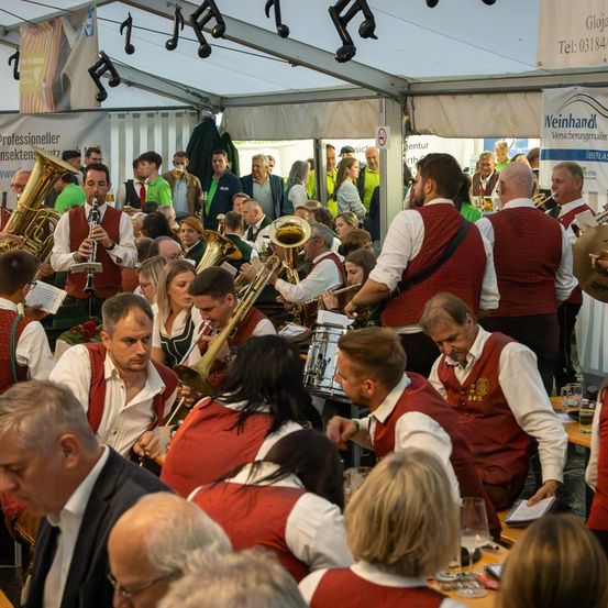 Eine Blaskapelle spielt in einem Zelt mit einem großen Publikum. Musiker spielen verschiedene Instrumente, und einige Zuschauer halten Getränke. Werbung ist im Hintergrund sichtbar.