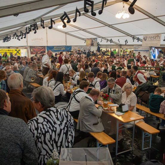 Eine große Menschenmenge sitzt in einem Zelt an Tischen, einige tragen traditionelle Kleidung. Musiknoten und Banner schmücken das Zelt. Ein Mann in einem weißen Hemd steht in der Nähe eines Tisches mit Essen.